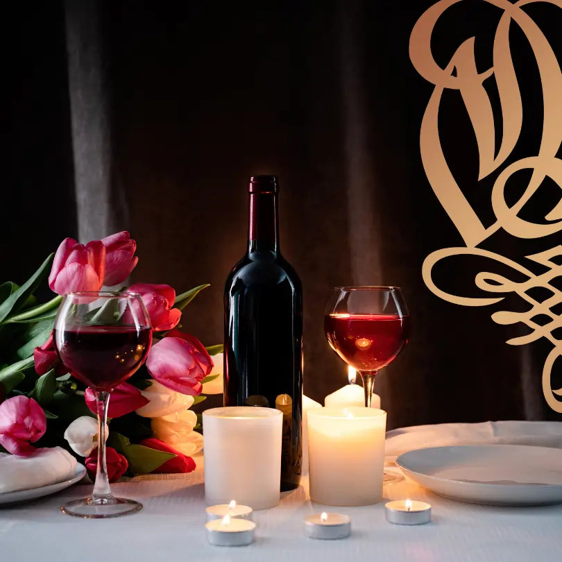 Candlelight Dinner Wöltingerode. Flasche Wein und ein Glas Wein stehen hinter einer Kerze. Das Wöltingerode W ist zur Hälfte zu sehen. Auf der linken Seite stehen Blumen